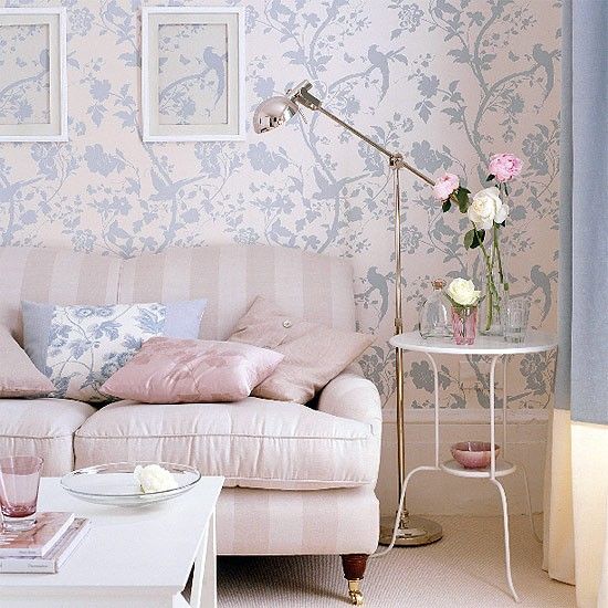 Sala de estar pequena, sofá rosa-claro com almofadas em tons de rosa e azul e mesa de canto com objetos decorativos.
